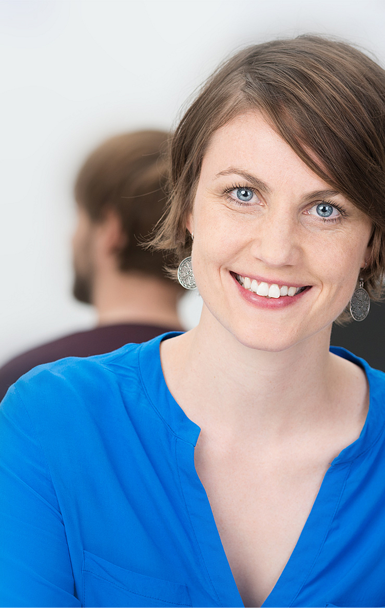 Eine lächelnde Frau mit kurzen, braunen Haaren trägt ein blaues Hemd. Sie sitzt in einem modernen Büro und schaut direkt in die Kamera. Im Hintergrund sind unscharf zwei weitere Personen bei der Arbeit zu sehen.
